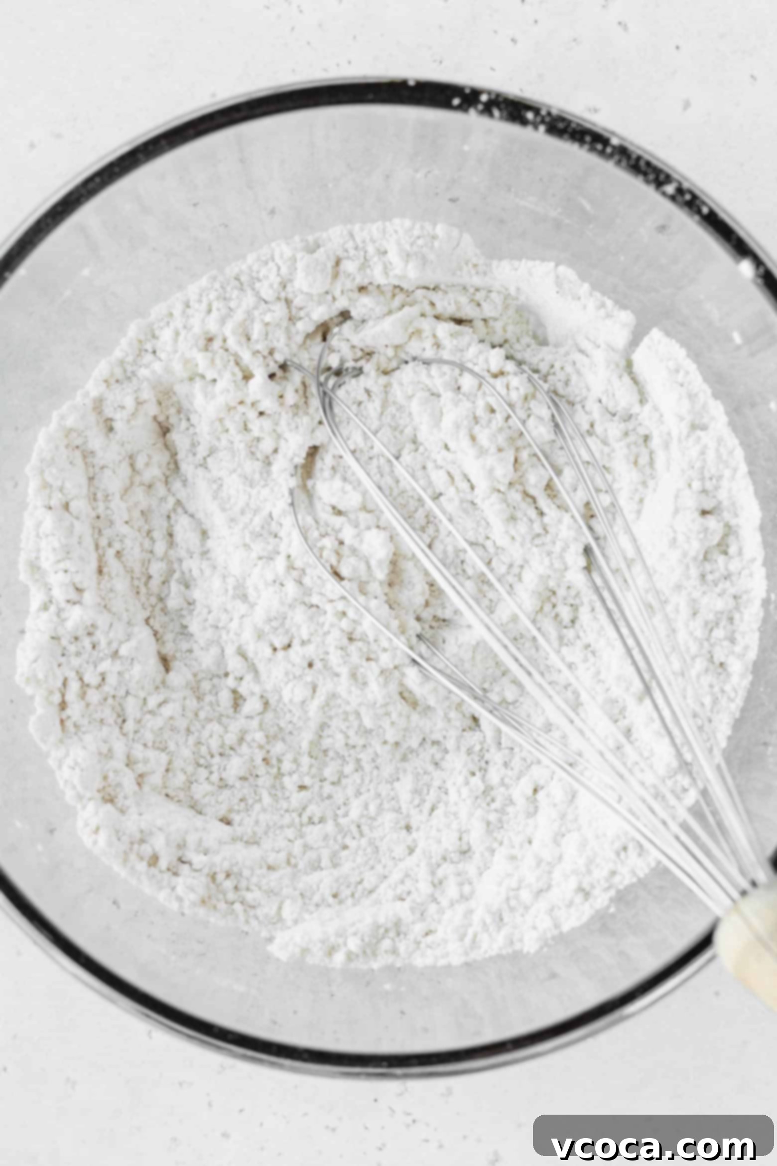 A bowl of dry ingredients including flour, salt, baking soda, and baking powder, with a whisk ready to combine them.