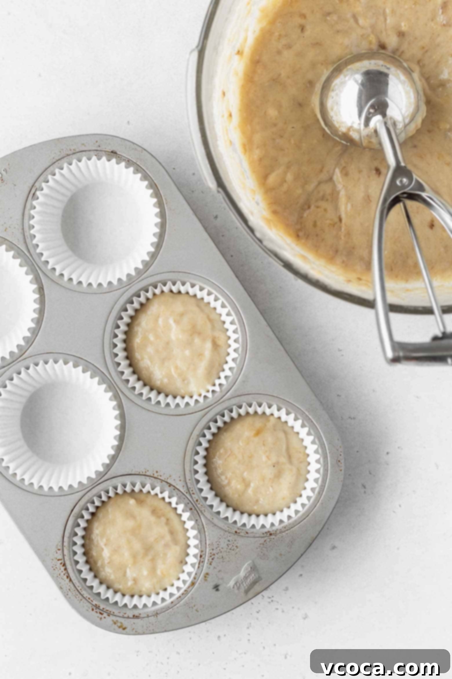Vegan banana cupcake batter being scooped into paper-lined cupcake pan using an ice cream scoop, ensuring even portions.