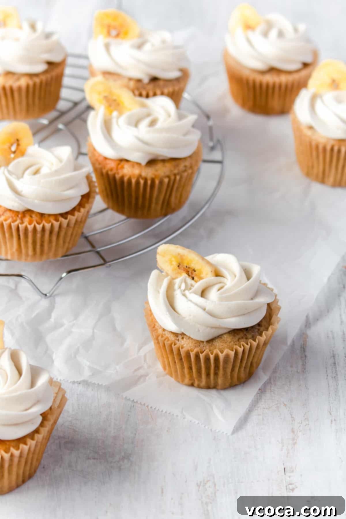 Beautifully frosted banana cupcakes with buttercream and a banana chip garnish, elegantly arranged on a white surface.