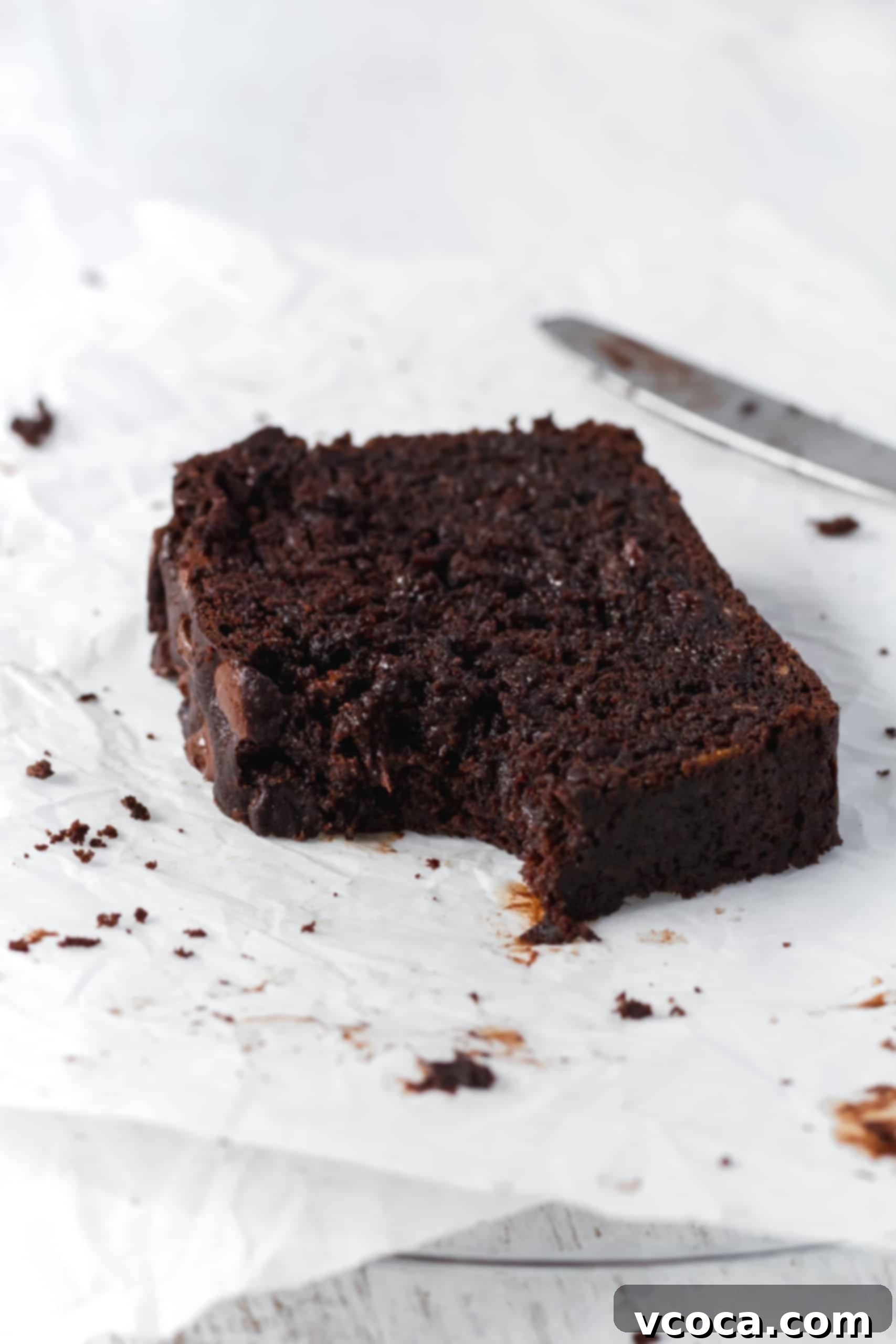Decadent Chocolate Banana Loaf 4 A close-up of a bitten slice of moist double chocolate banana bread, resting on parchment paper, showing the rich chocolate color, soft texture, and delectable melted chocolate pools inside. It looks incredibly appetizing.