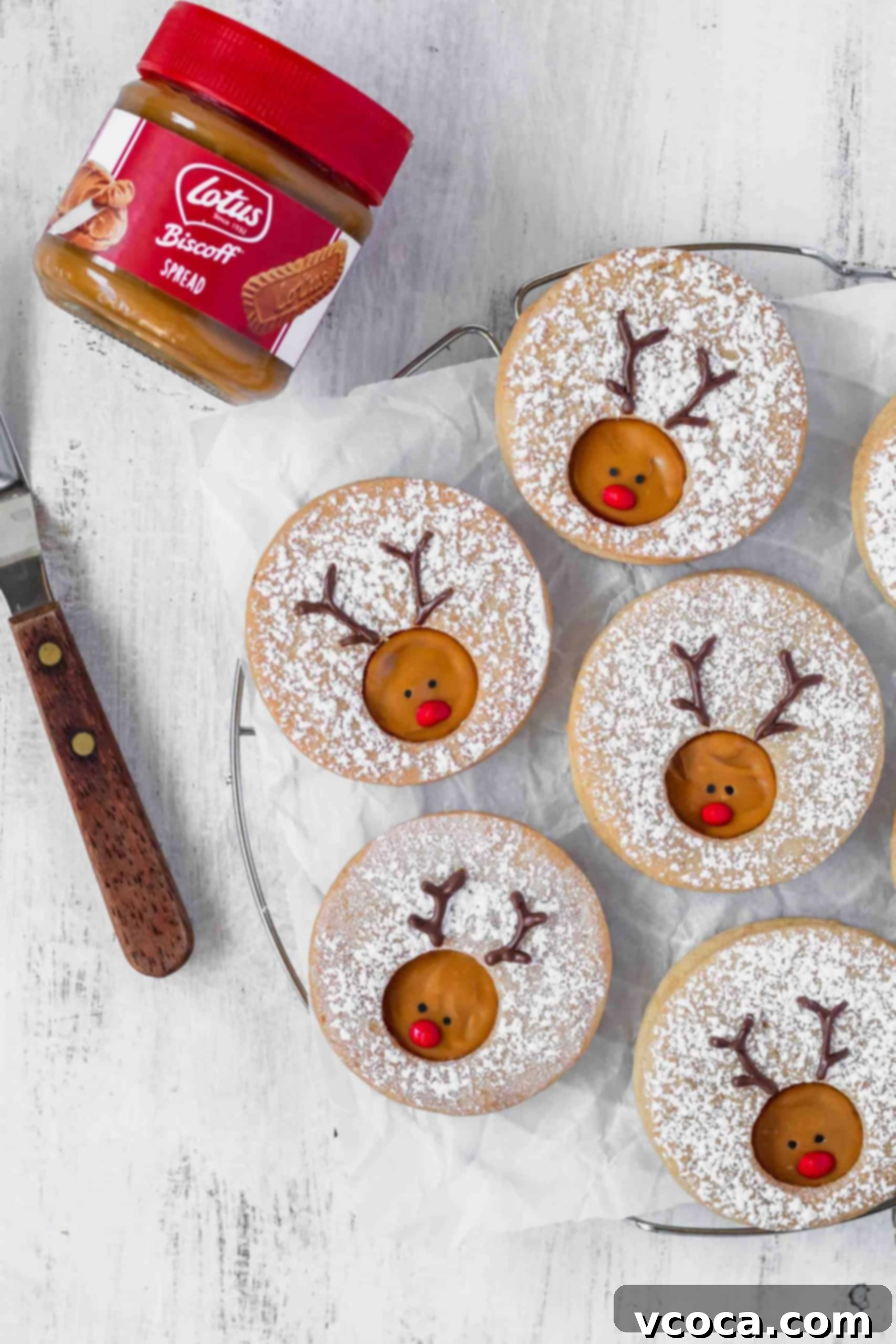 Rudolph's Holiday Reindeer Cookies 3 Reindeer cookies laying on a piece of parchment paper next to a jar of Biscoff spread.