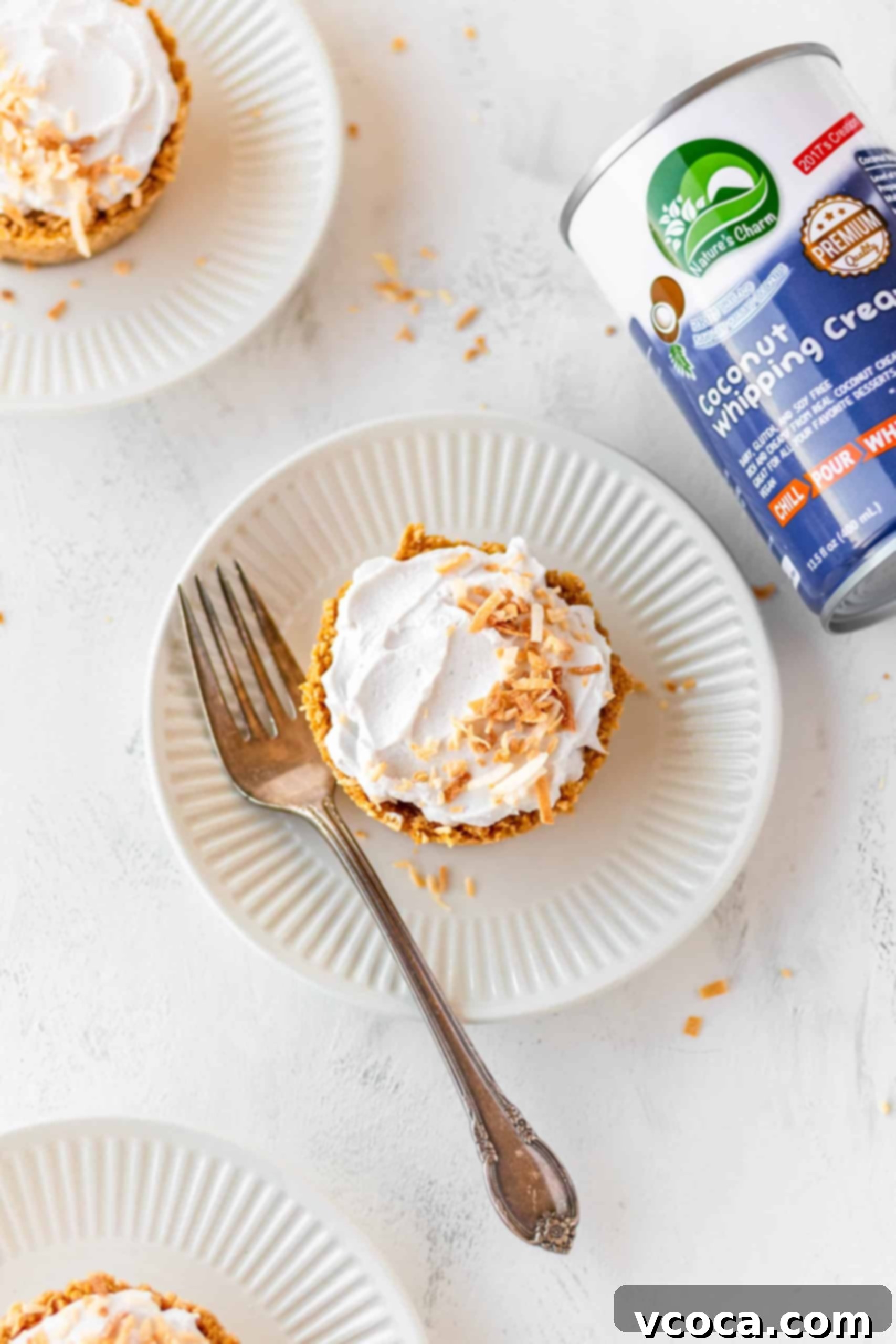 Mini Vegan & Gluten-Free Coconut Cream Pies 4 A single vegan coconut cream pie on a white plate with a fork, showing the distinct layers of crust, custard, and whipped cream.
