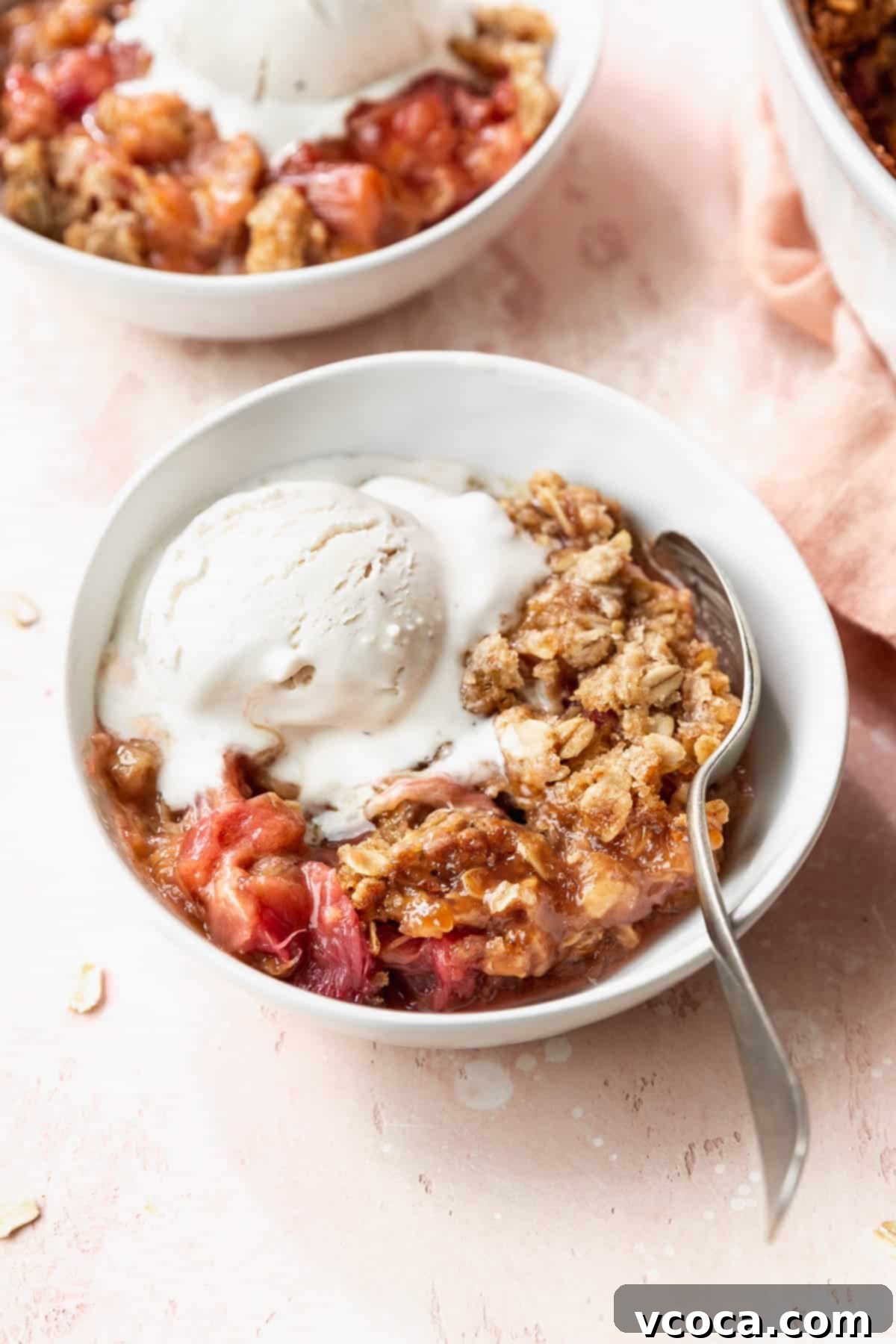 Sweet & Tart Gluten-Free Rhubarb Crisp 9 45 degree angle shot of a white bowl filled with gluten-free rhubarb crisp with oats crumble topping and a scoop of vanilla ice cream on a pink table.