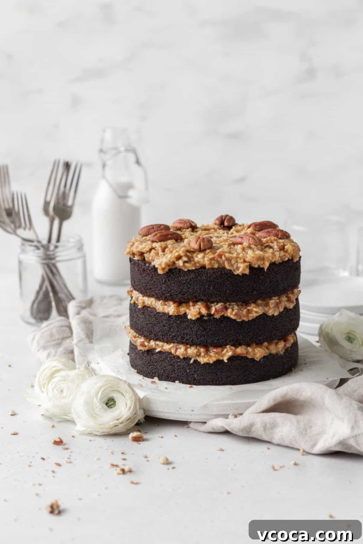 Decadent Gluten-Free German Chocolate Dream 2 A three-layer gluten-free German chocolate cake on a round wooden board with white flowers beside it, with a milk jug and a jar with forks inside in the background.