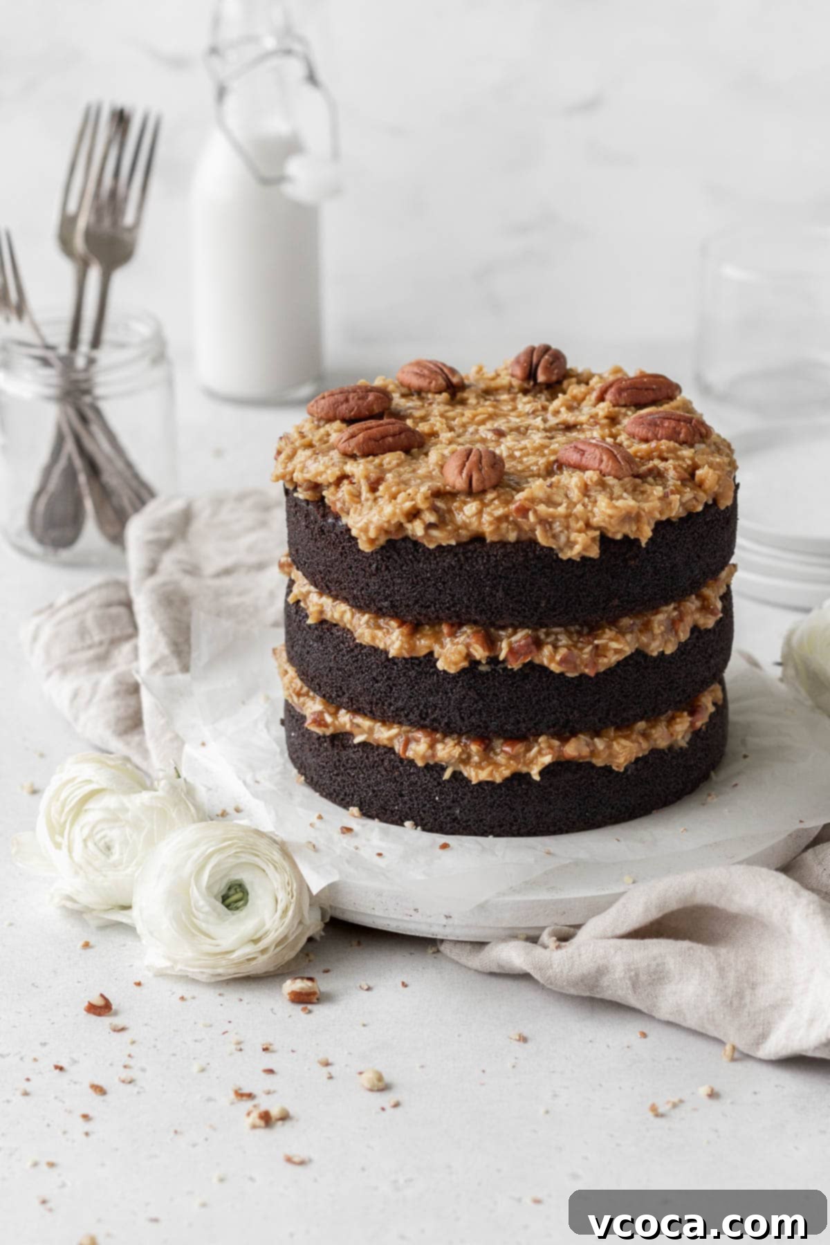 Decadent Gluten-Free German Chocolate Dream 15 A three-layer German chocolate cake on a round white wooden board next to white flowers.