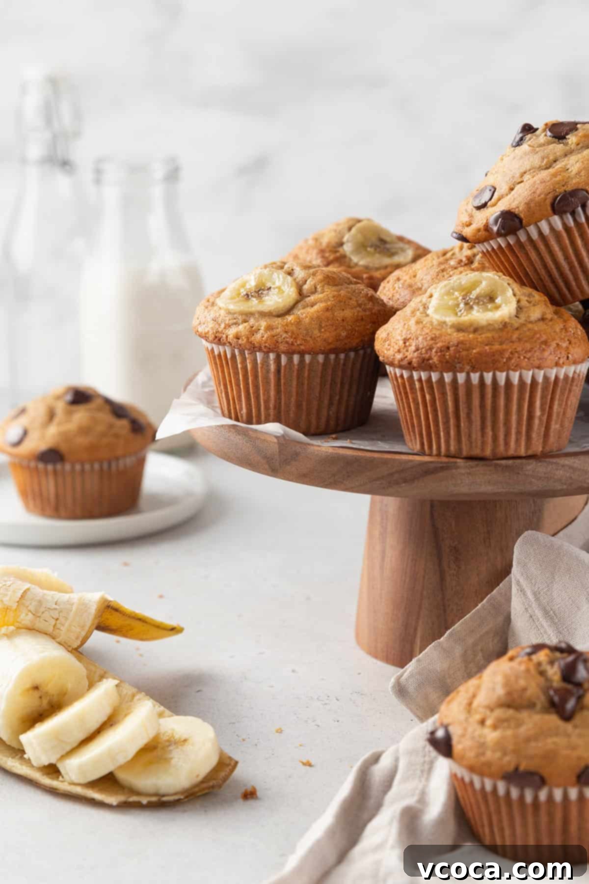 Irresistible Gluten-Free Banana Muffins 2 Gluten-free banana muffins on a wooden cake stand, with some sliced bananas on the surface underneath, and glass milk bottles in the background.