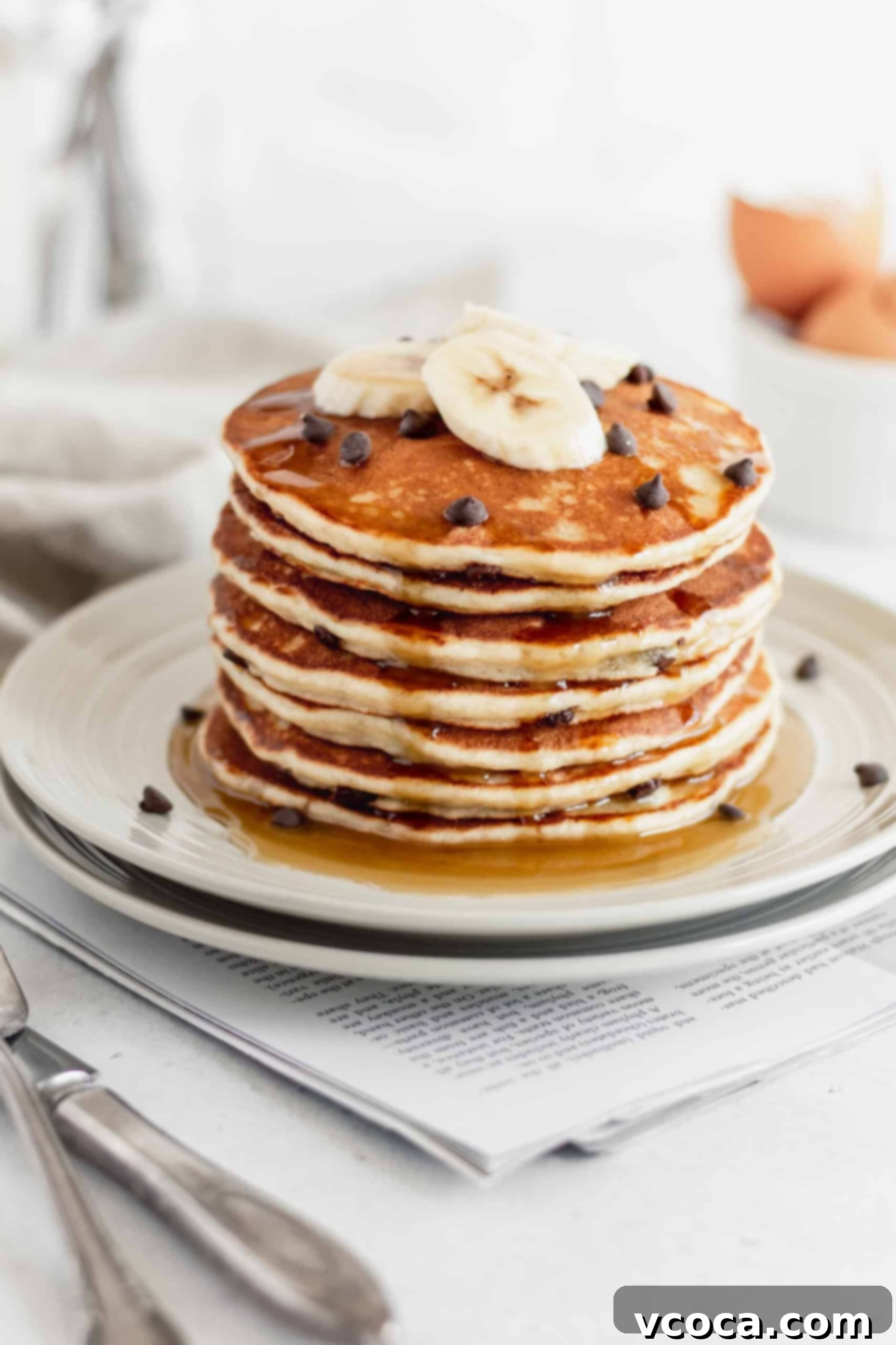 Fluffy Gluten-Free Banana Pancakes 2 A tall stack of fluffy gluten-free banana pancakes on a white plate, topped with fresh banana slices and a light dusting of powdered sugar. A generous drizzle of golden maple syrup cascades down the sides, highlighting their moist and tender texture.