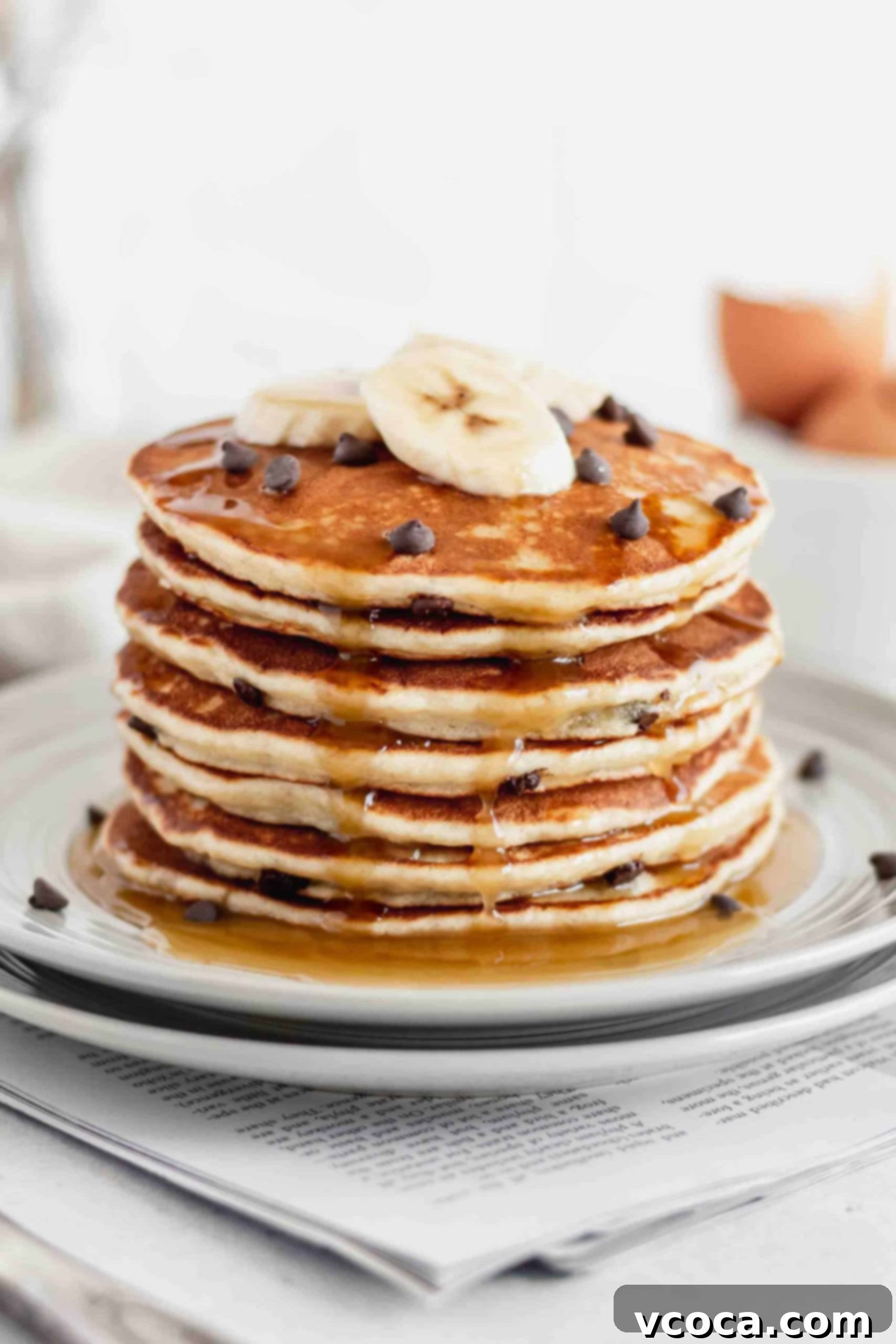 Fluffy Gluten-Free Banana Pancakes 4 A glorious stack of gluten-free banana chocolate chip pancakes generously drizzled with rich, golden maple syrup. Slices of fresh banana and scattered chocolate chips adorn the top, enhancing the visual appeal and suggesting a delicious indulgence.