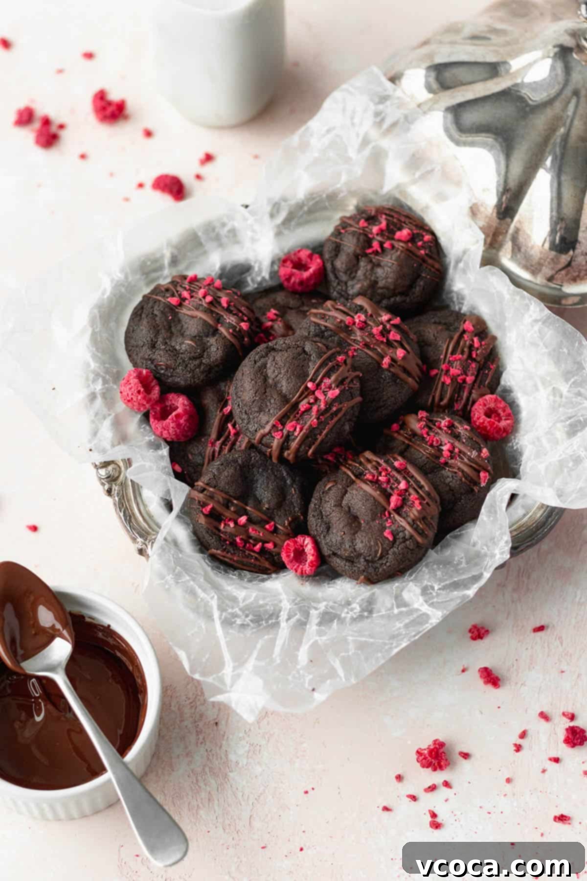 Overhead shot of double chocolate chip cookies in a silver dish lined with parchment paper, with freeze-dried raspberries sprinkled all around. The cookies are perfectly round and rich.