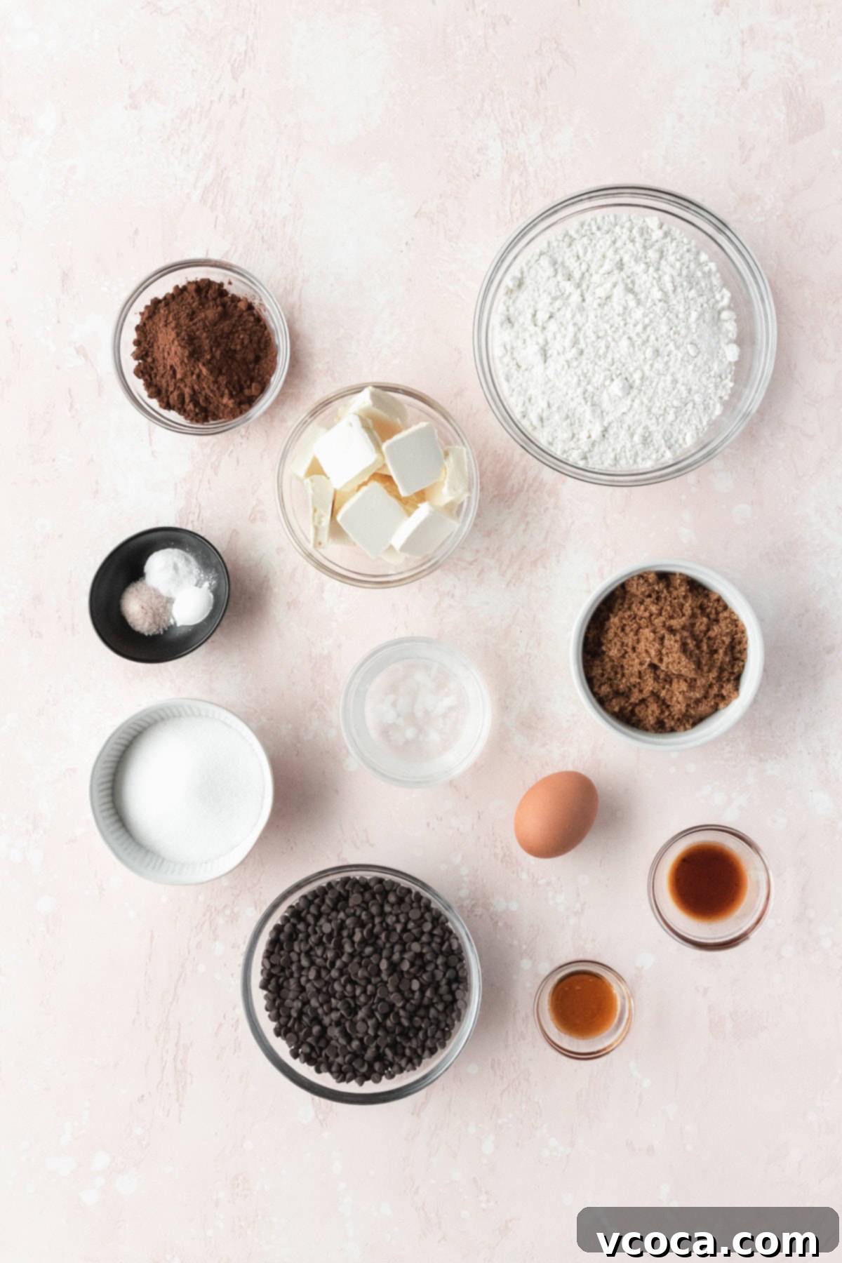 Ingredients to make double chocolate chip cookies measured out into small bowls and laid out on a pink surface. Various bowls of flour, cocoa, sugars, butter, egg, and chocolate chips are neatly arranged, ready for baking.