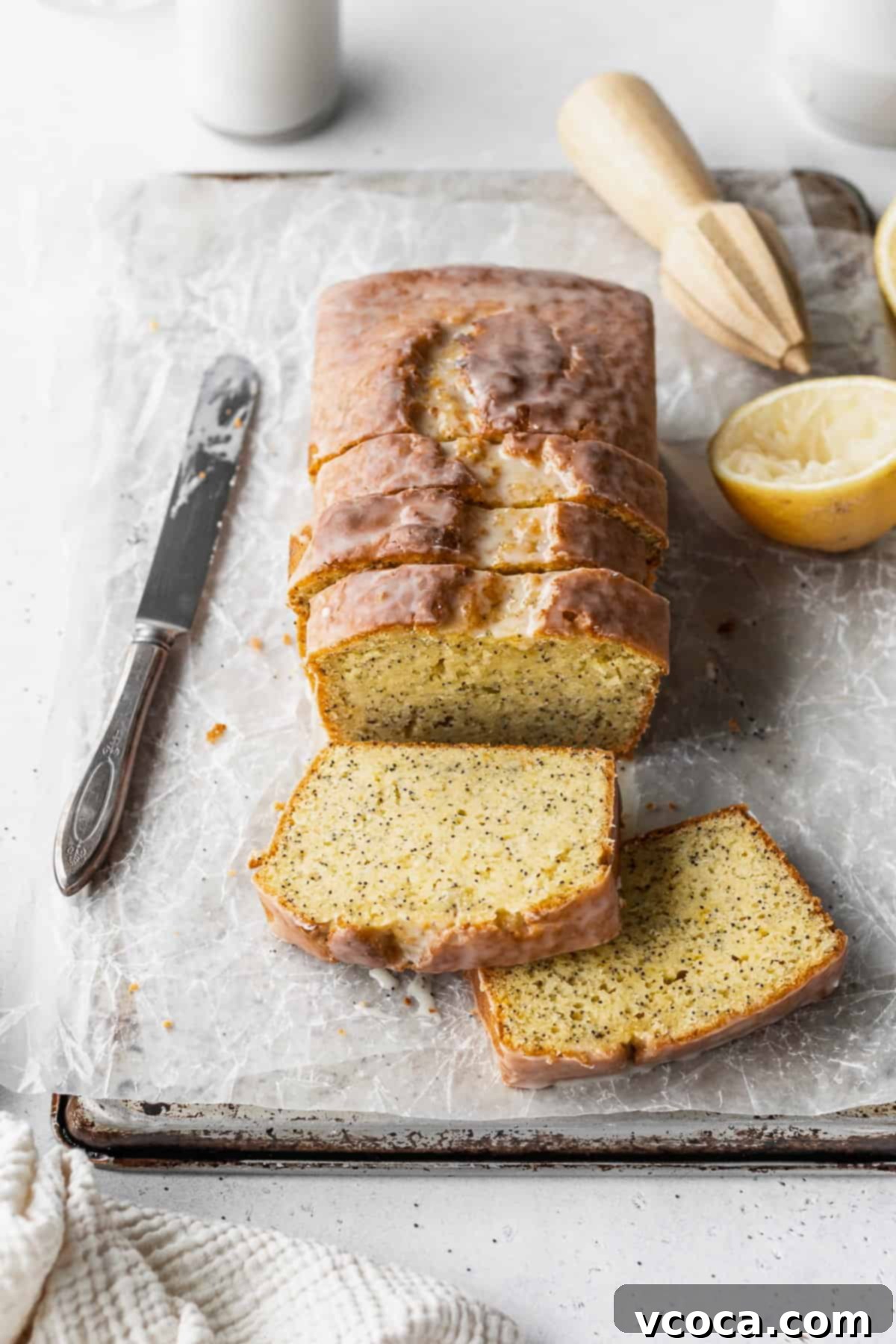 Zesty Gluten-Free Lemon Poppy Seed Cake 2 A sliced loaf of gluten-free lemon poppy seed loaf on a parchment paper lined baking sheet with a knife, half a lemon, and a wooden citrus zester placed around it.