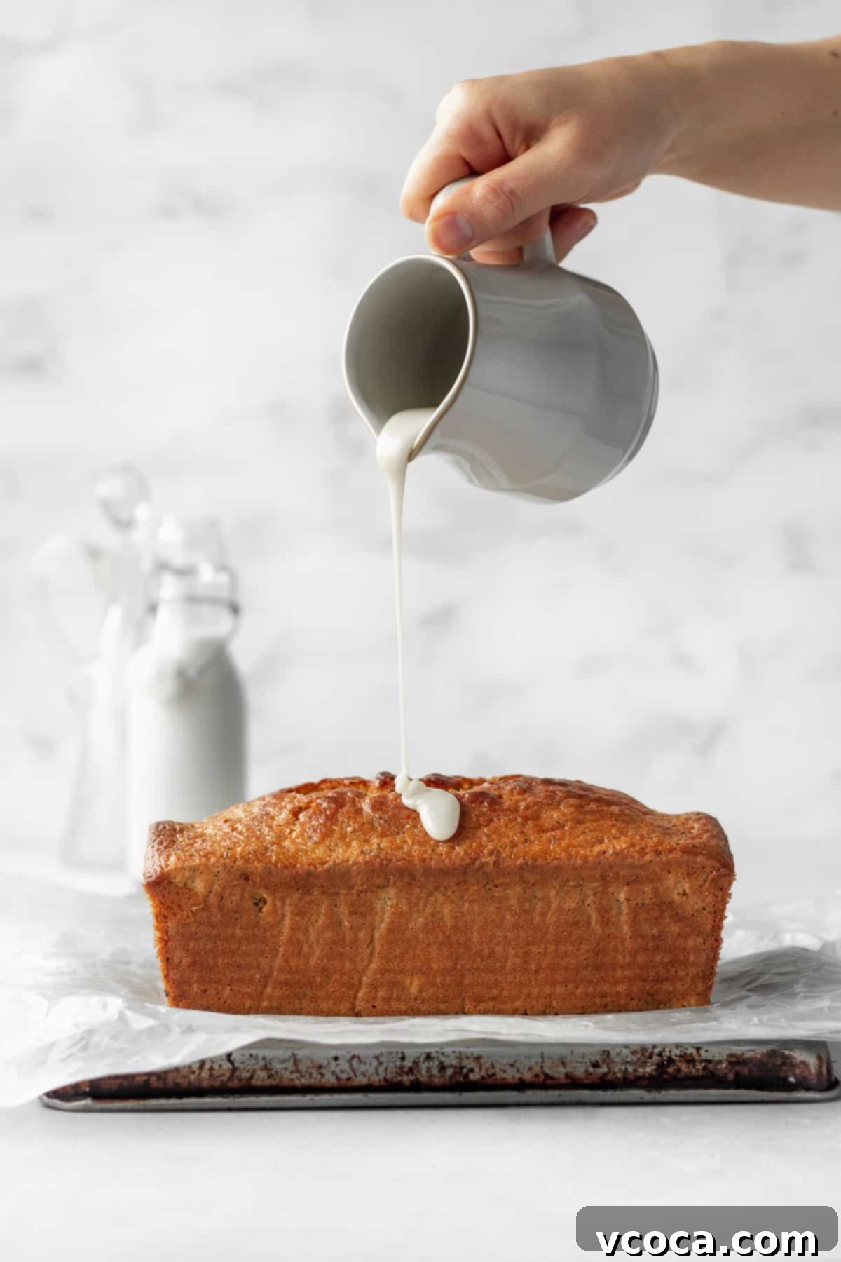 Zesty Gluten-Free Lemon Poppy Seed Cake 4 A hand holding a ceramic jug of lemon icing being poured onto a loaf of gluten free lemon poppy seed cake.