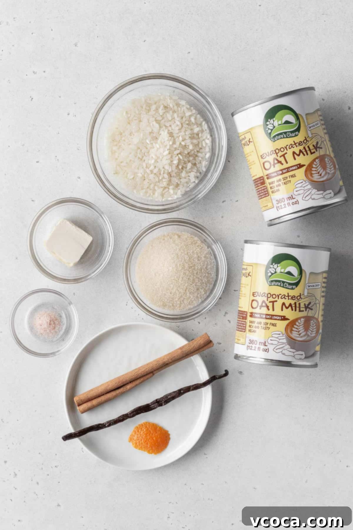 Dreamy Vegan Rice Pudding 3 Vegan rice pudding ingredients measured out into bowls with 2 cans of evaporated oat milk on a white surface.