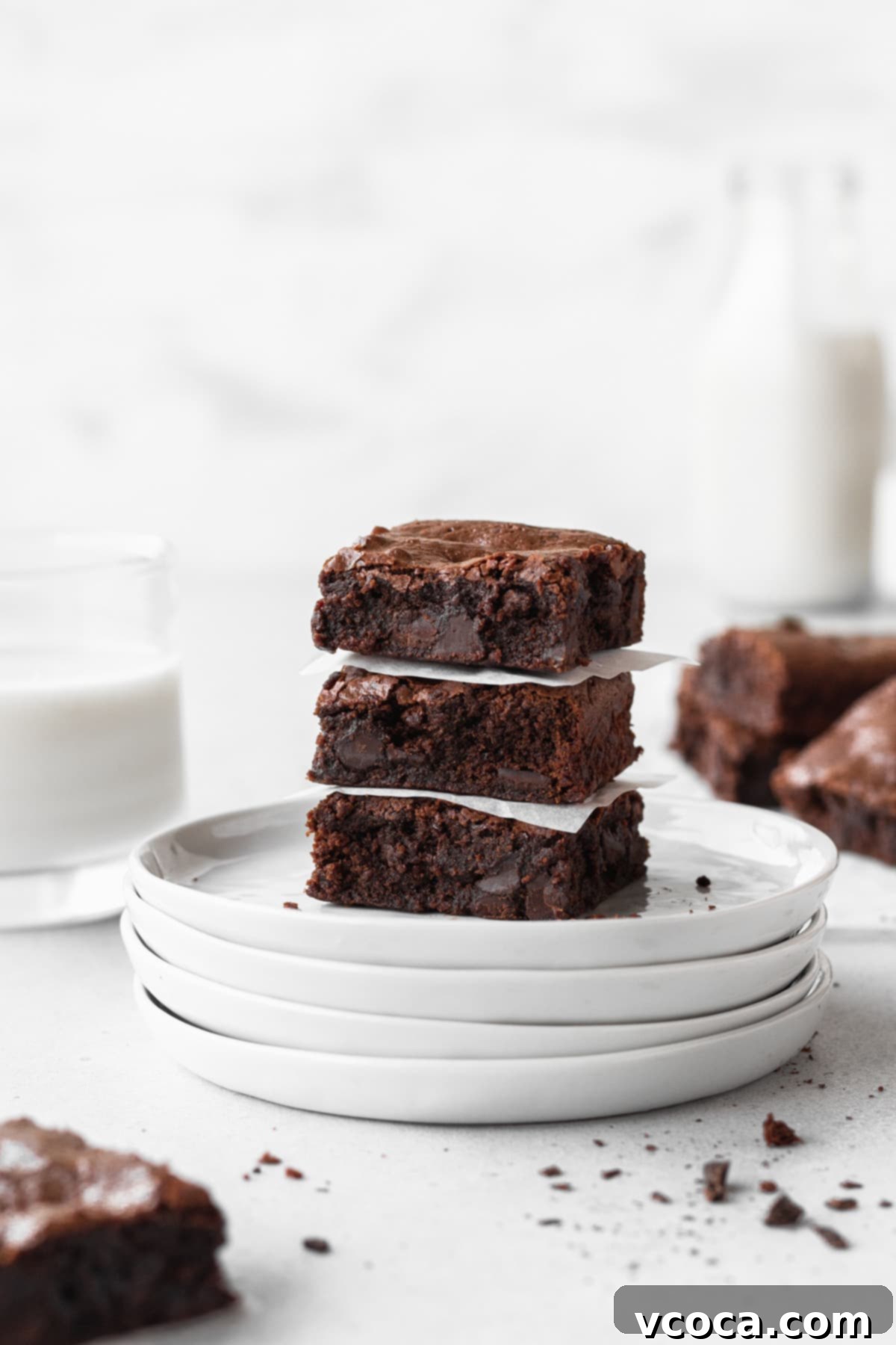 Decadent Gluten-Free Brownies 10 Three perfectly stacked gluten-free chocolate brownies on white plates, with more brownies and a jug of milk blurred in the background, highlighting their rich color and texture.